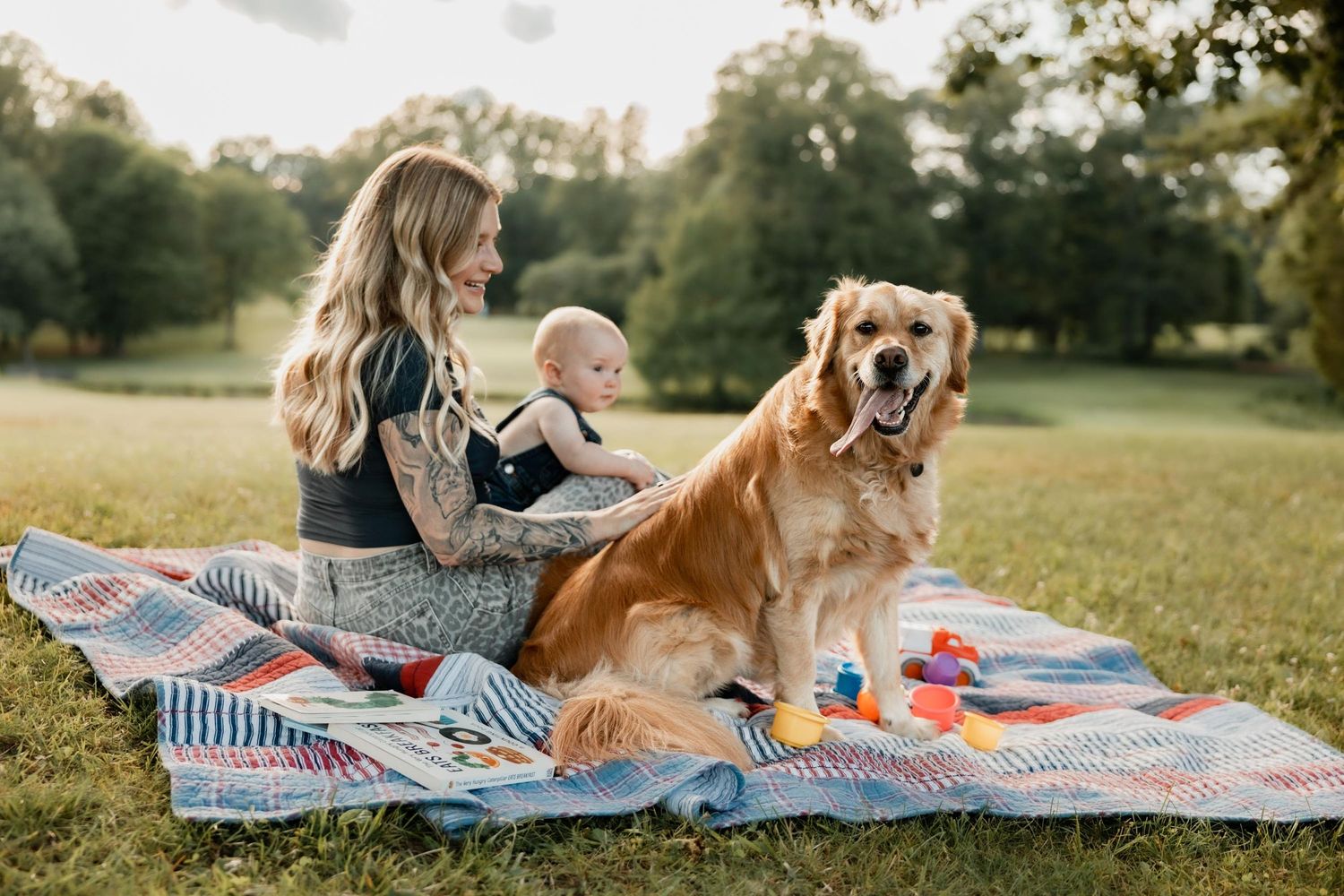 A woman with tattoos sits on a colorful quilt in a park with a baby and a golden retriever dog.