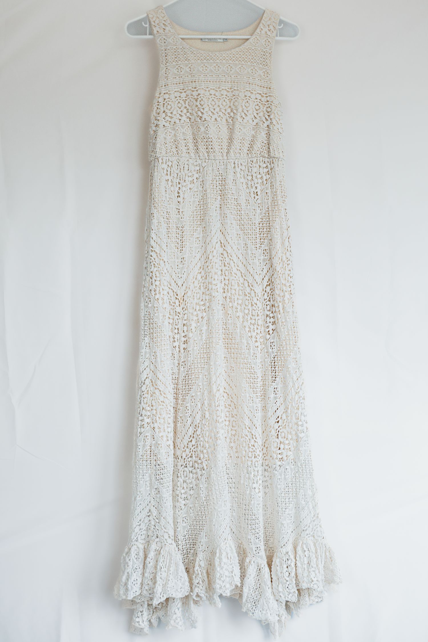 Vintage white lace sleeveless summer dress with delicate crochet pattern and ruffled hem displayed on hanger against wall.