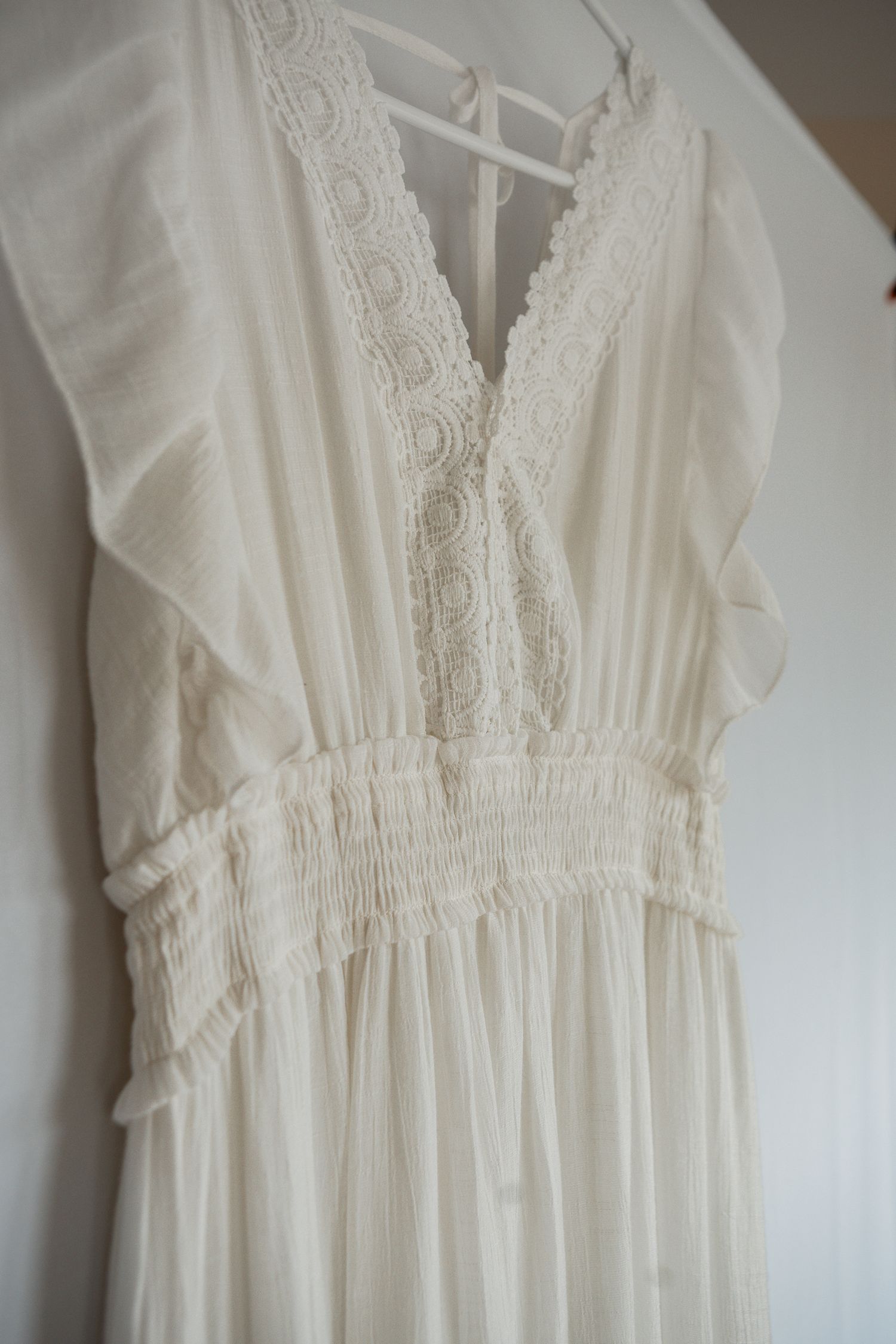 Delicate white lace Victorian-style dress with v-neckline and gathered empire waist displayed against a light wall.