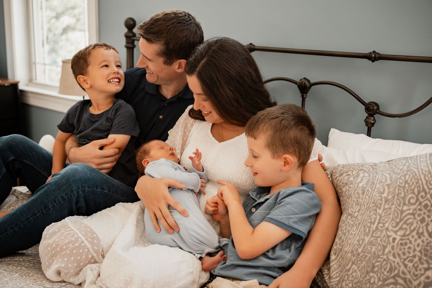 Family relaxes together on grey couch while looking at electronic device in bright living room.