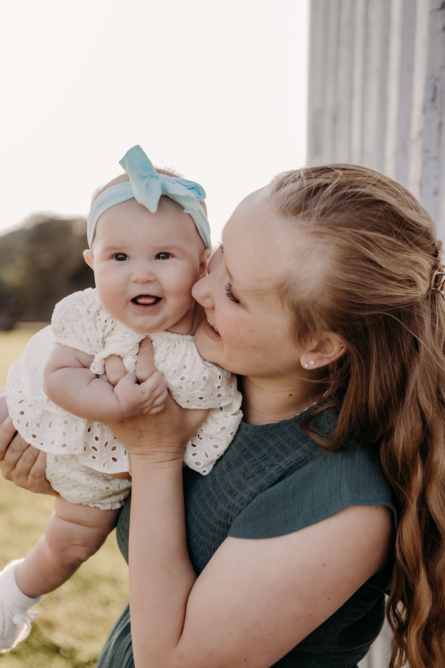 A tender moment outdoors as a woman in a dark top lovingly holds a baby wearing a light dress and blue bow headband.