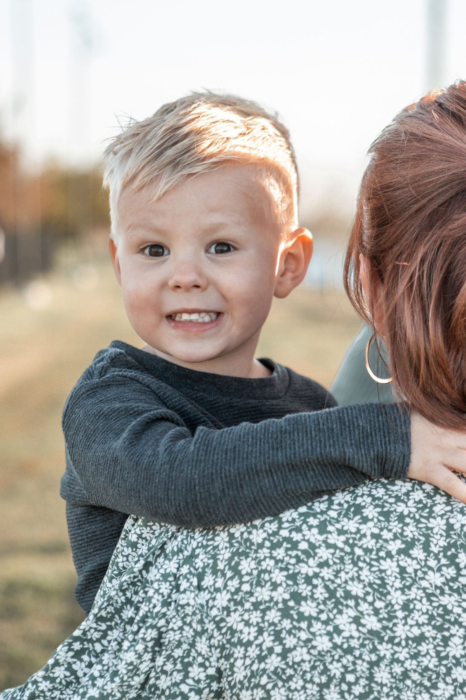 A happy toddler in a dark sweater hugging someone wearing a floral dress in warm sunlight.