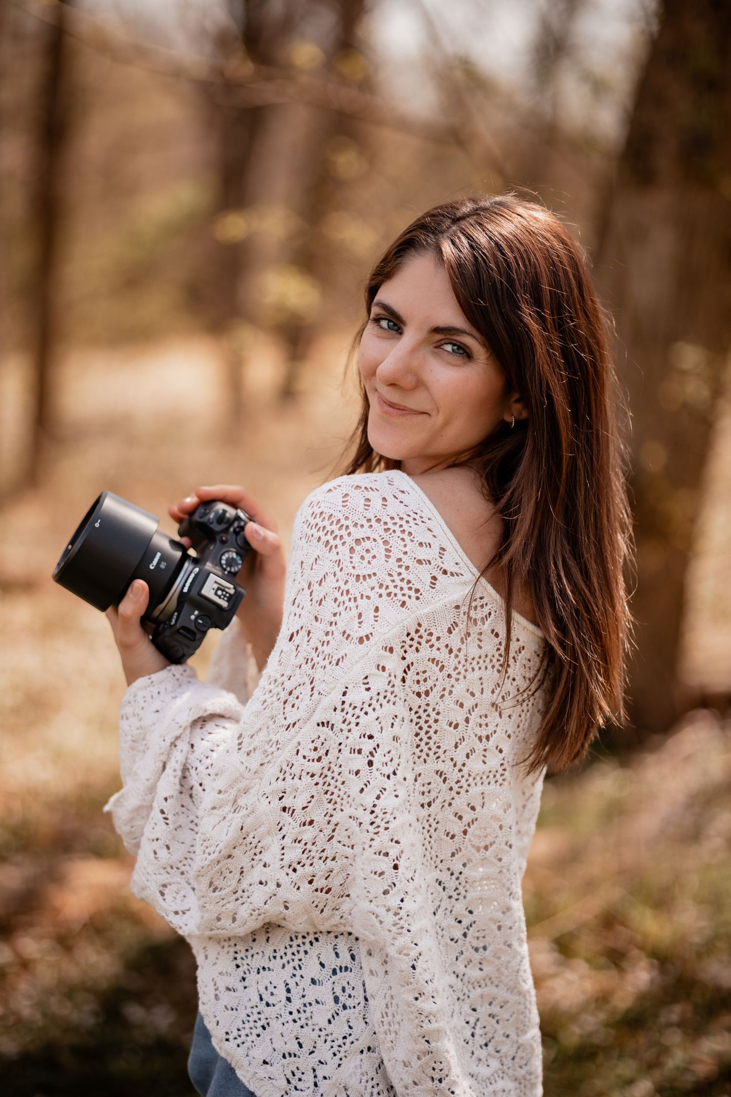A smiling brunette woman in a white lace sweater holds a DSLR camera outdoors in an autumn forest setting.