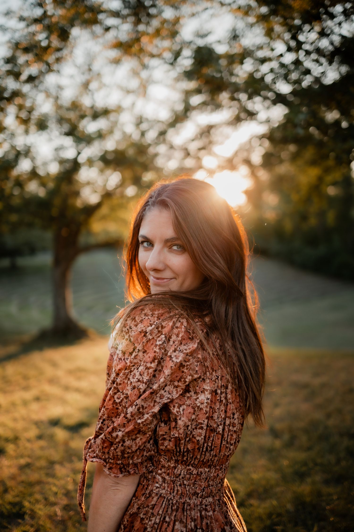 Woman in floral dress smiling over shoulder at golden hour sunset in lush green park.