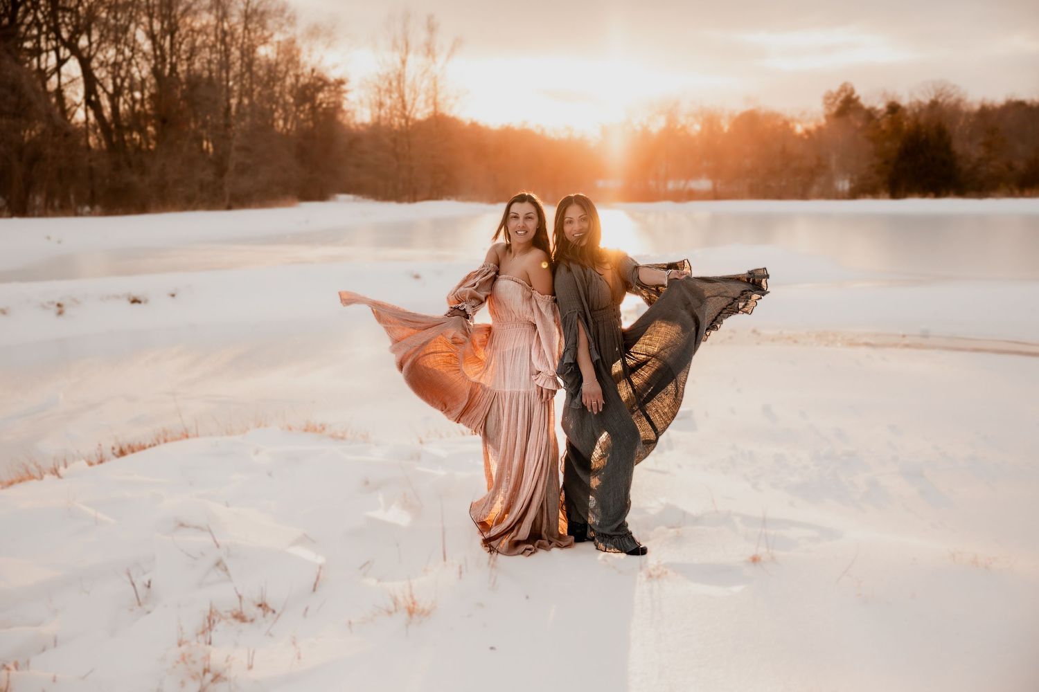 Two women in flowing gowns pose in snowy landscape at golden sunset, dresses billowing in the wind.Lifestyle Virginia Photographer Anglin