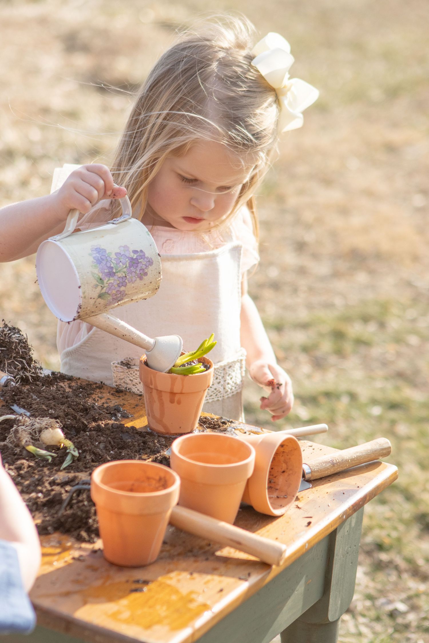 Child wearing white bow carefully waters small terracotta pots on wooden table outdoors.
