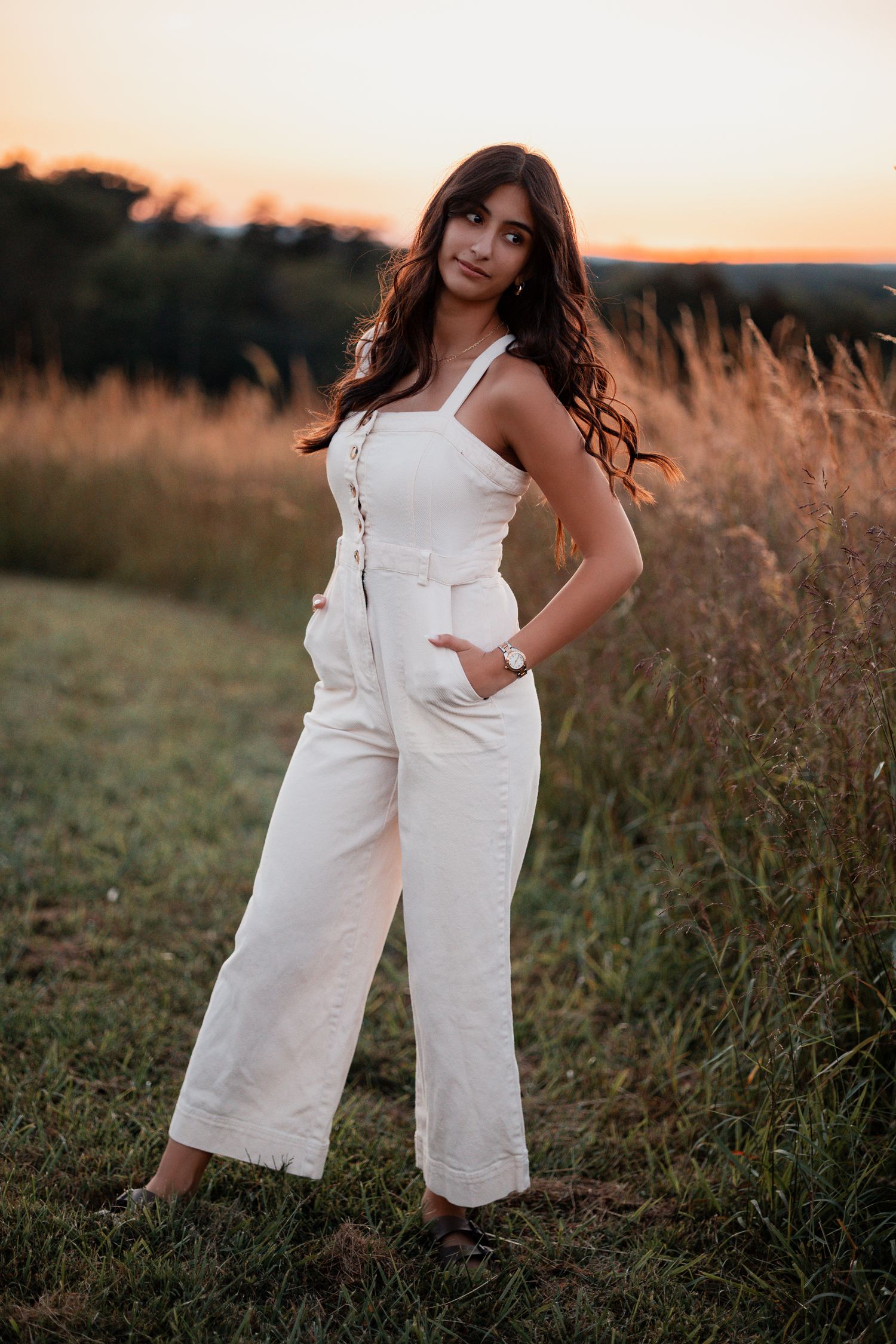 A woman poses in a white jumpsuit against a backdrop of tall grass during golden hour.