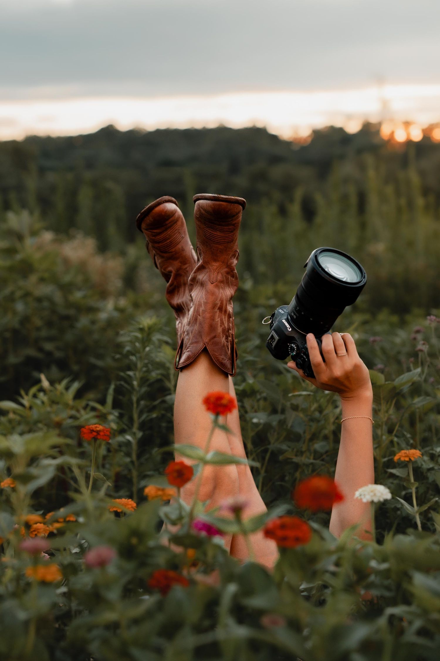 Legs with brown cowboy boots and an arm holding a camera raised above a colorful flower field at sunset.