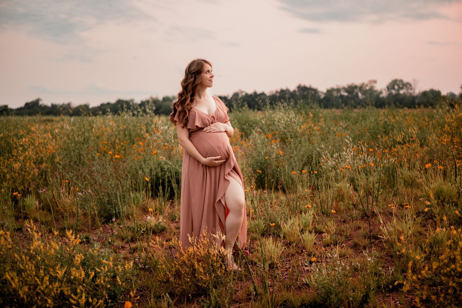 A pregnant woman in a flowing pink dress stands in a golden wildflower field during sunset.