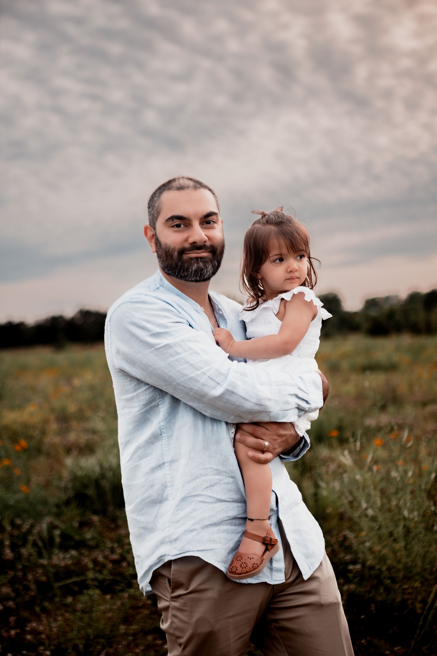 A father holds his young daughter in a field at sunset while wearing a light blue shirt and brown pants.