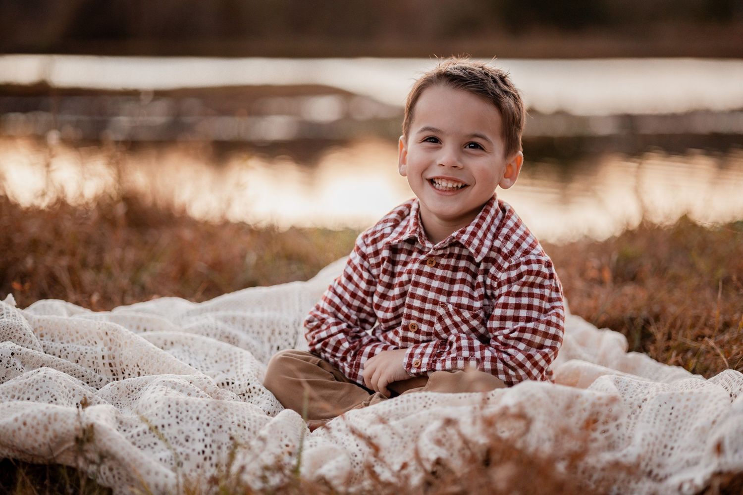 A child in a red and white checkered shirt sits on a textured white blanket during a golden hour outdoor photo session.
