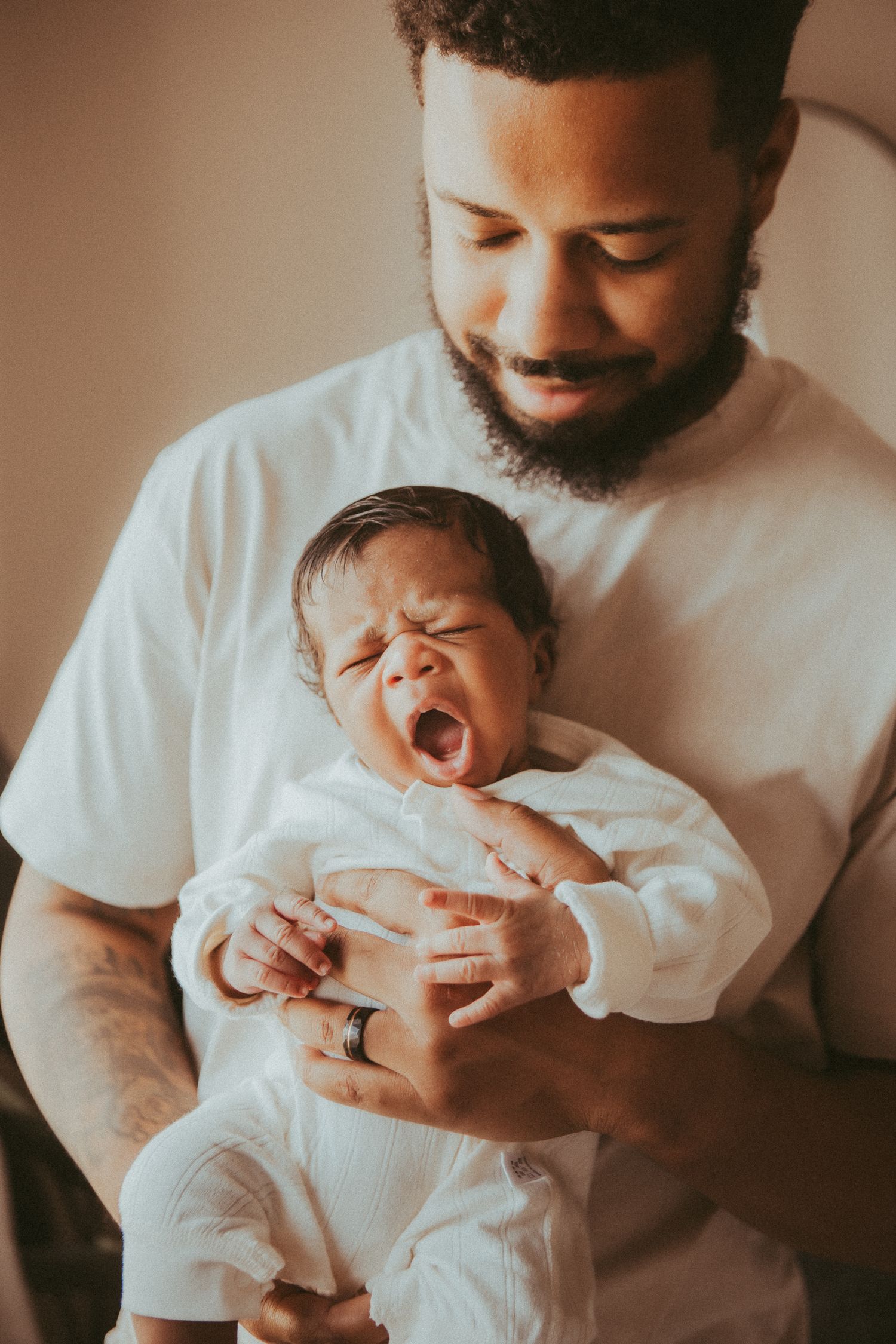 A tender moment shows a parent in a white t-shirt holding a yawning newborn baby dressed in white.