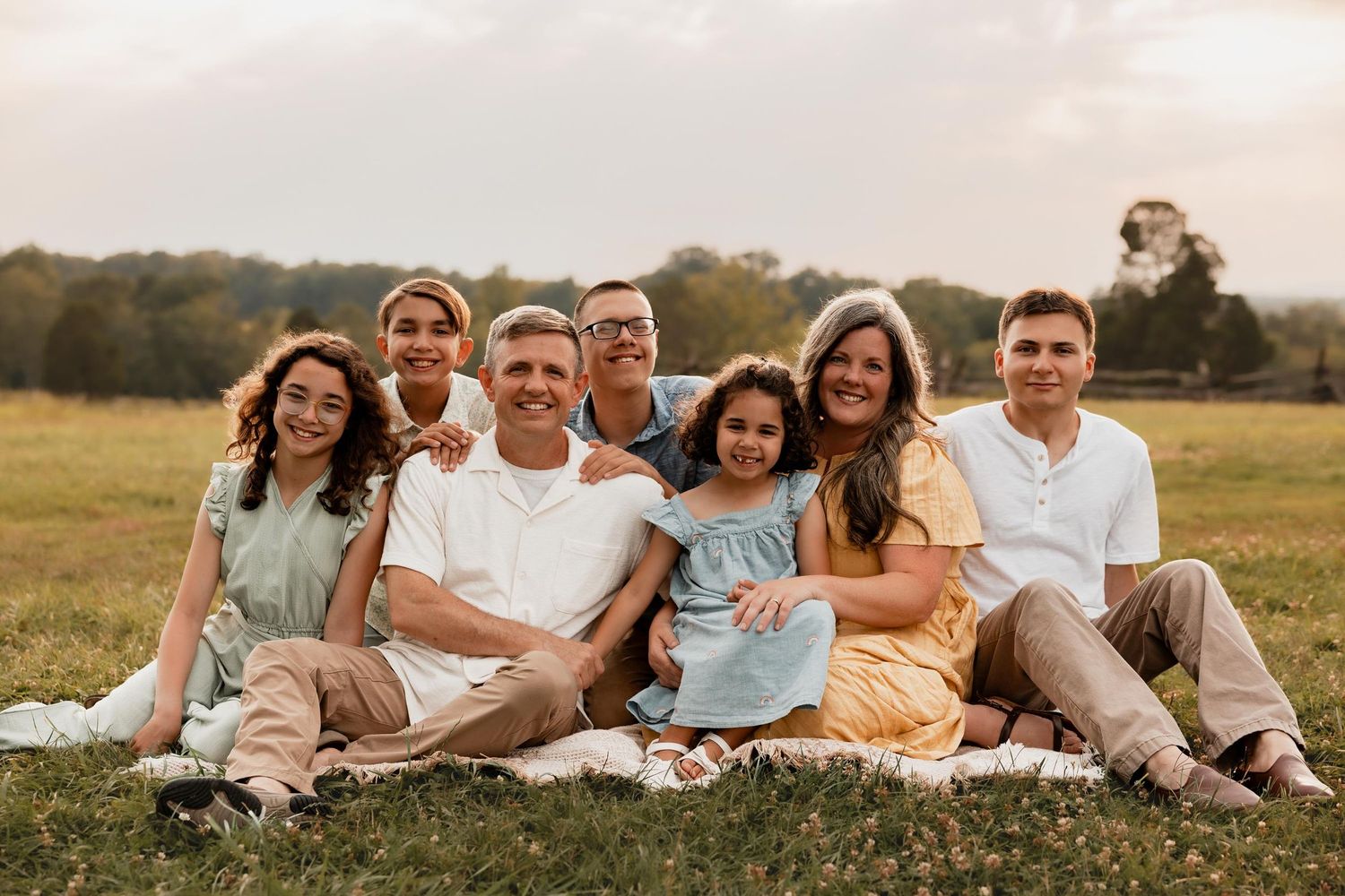 Happy family of seven sitting on a blanket in a green field, smiling together during a warm outdoor photo session.