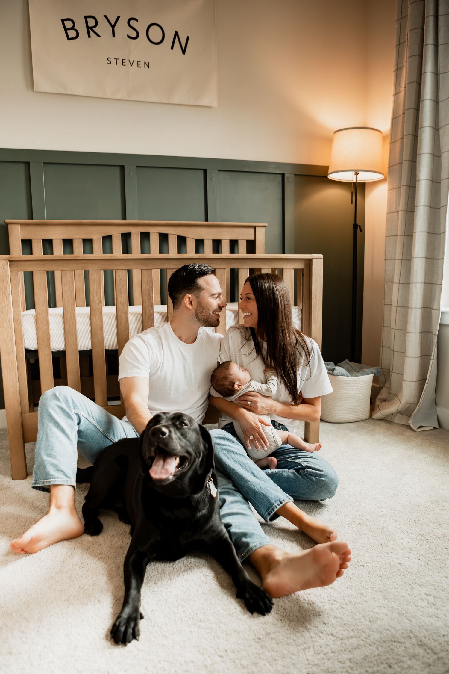 A couple and their black dog relax together on a carpeted floor in a cozy bedroom with soft lighting.