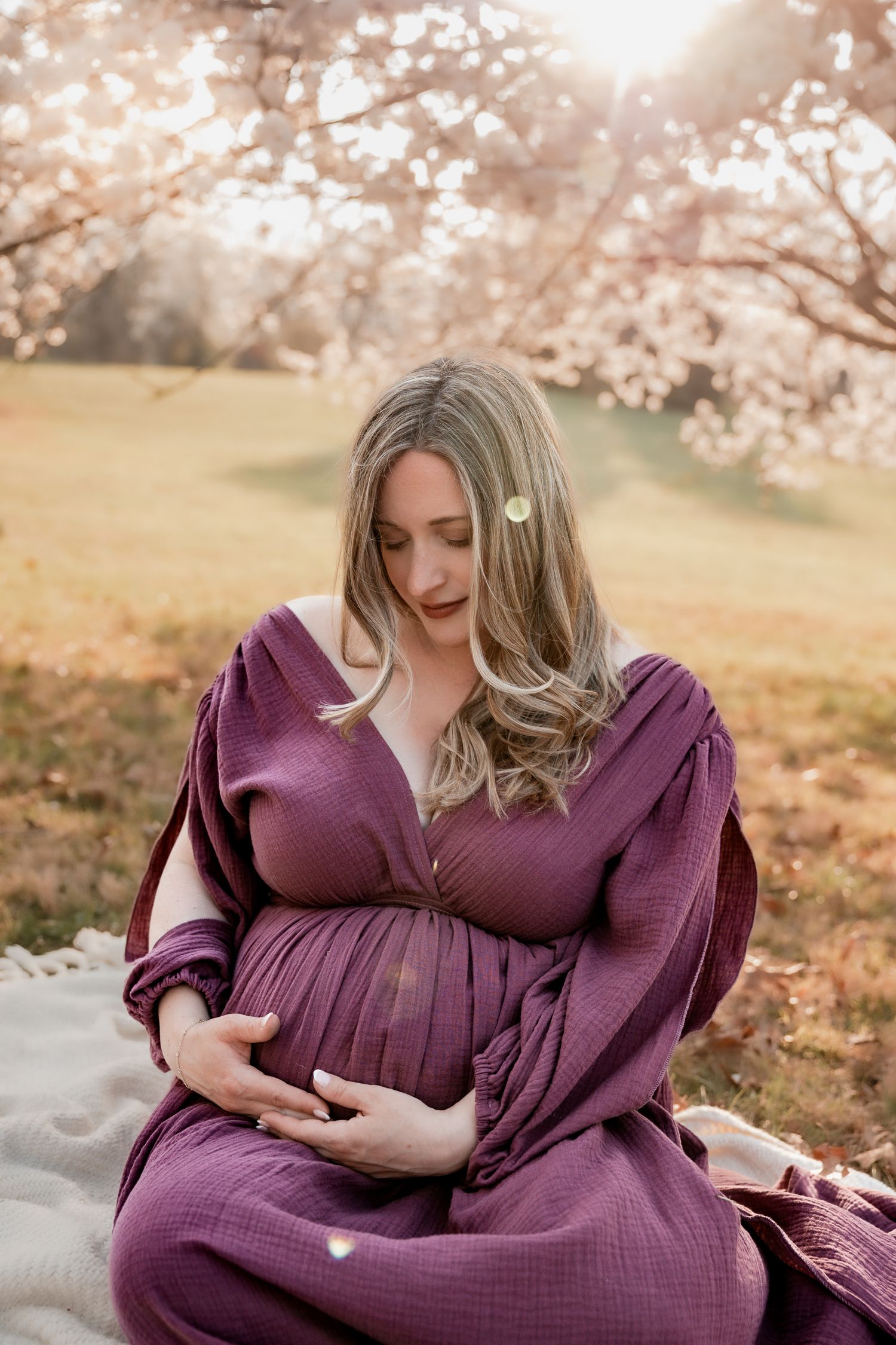 Pregnant woman in purple dress cradling baby bump outdoors under blooming cherry blossom trees at golden hour.
