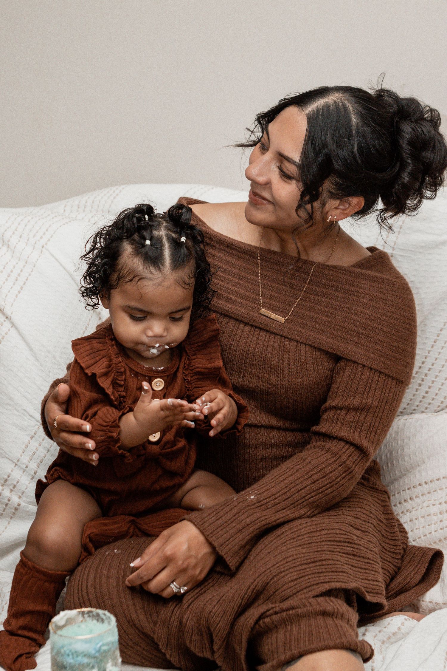 Mother and child wearing matching brown knit outfits sit together on a white couch in an intimate moment.