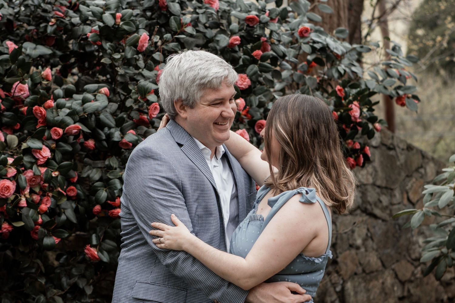 Happy couple embraces and laughs together in front of blooming red camellia bush in romantic garden setting.