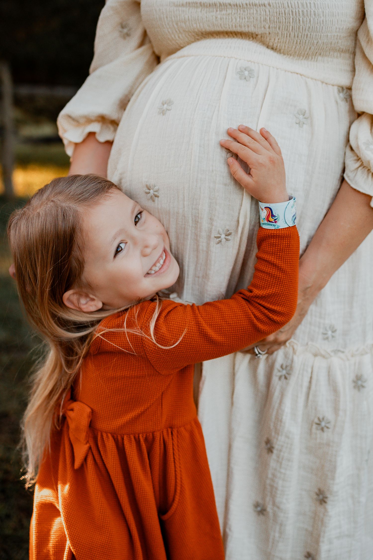Young girl in orange dress smiling while touching pregnant mother's belly outdoors.