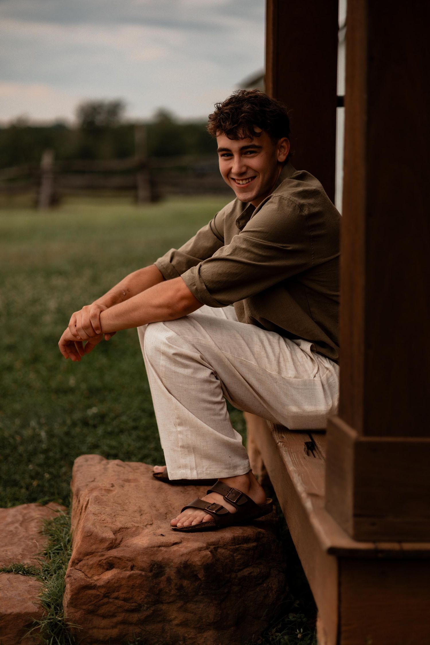 Smiling young man in olive shirt and white pants sitting on a rock ledge outside a rural building.