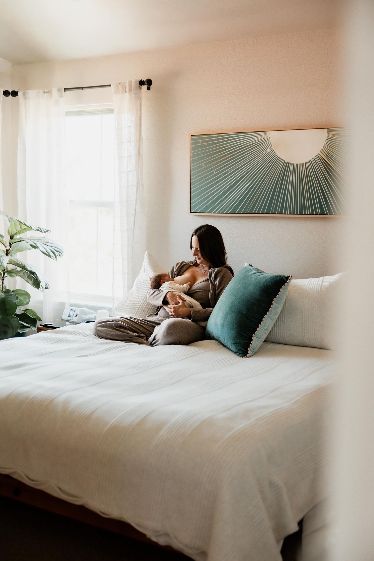 A mother sits cross-legged on a white bed nursing her newborn beneath a sunburst wall art piece.