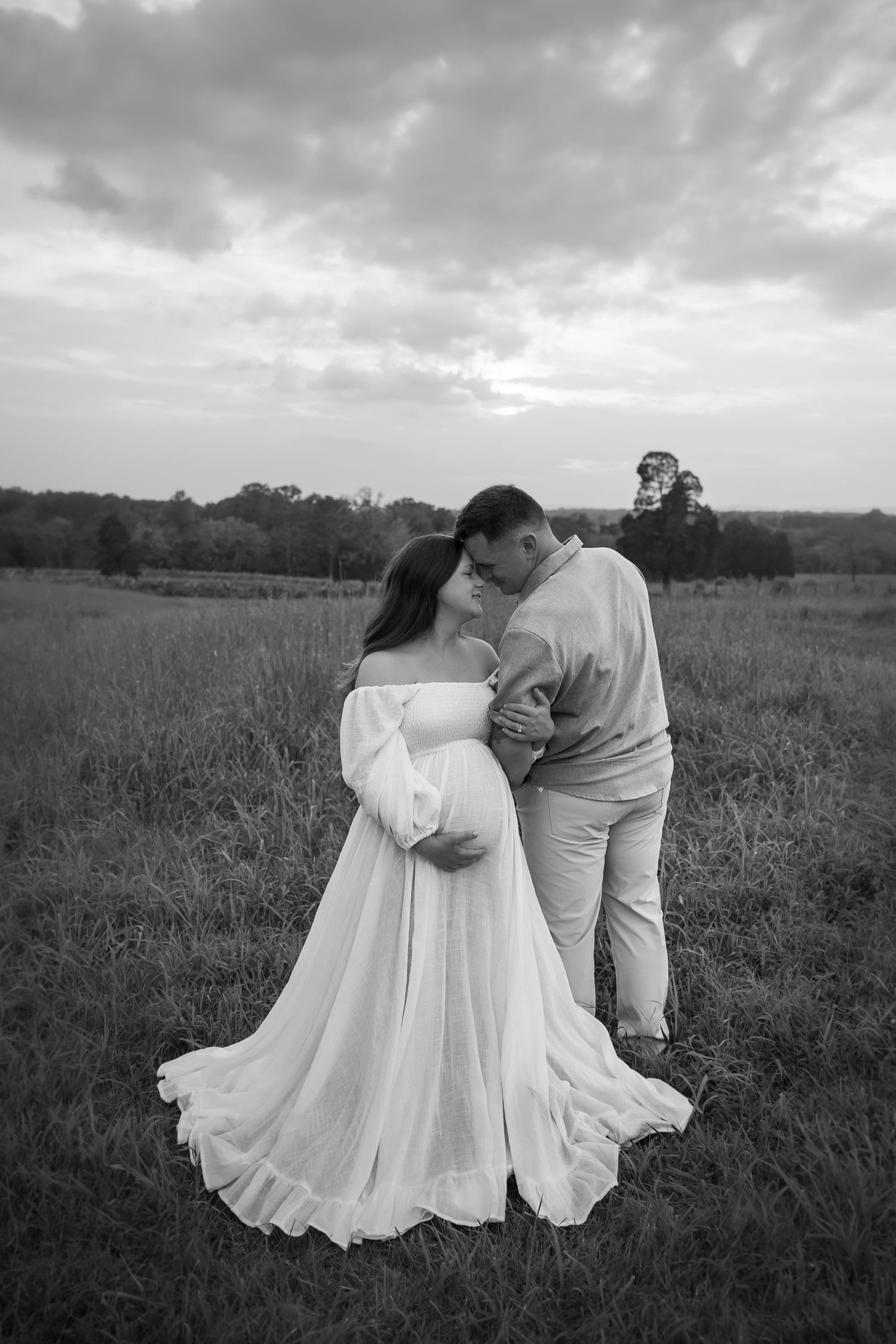 Couple sharing a kiss in open field at dusk, black and white maternity photo with woman in flowing white gown.