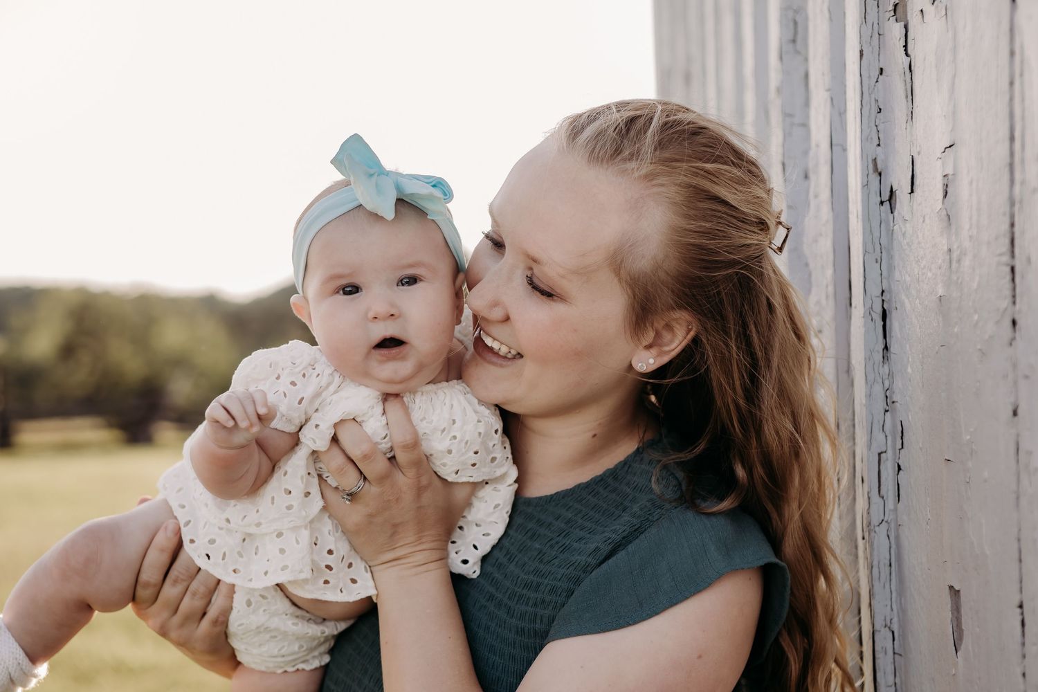 A tender outdoor portrait of a parent and baby in lace dress sharing a loving moment against white siding.