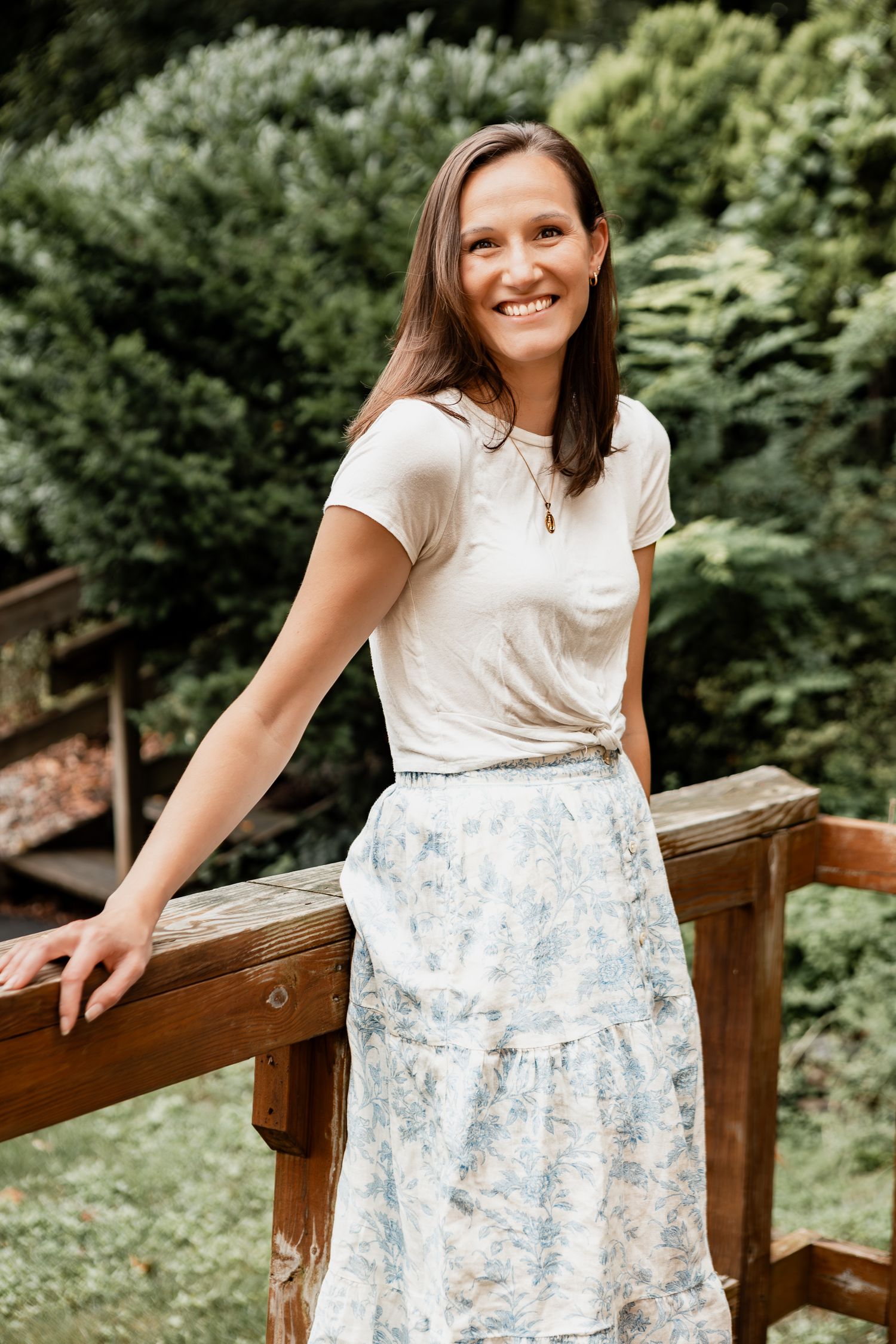Woman in white t-shirt and floral skirt poses outdoors on wooden deck railing with green foliage background.