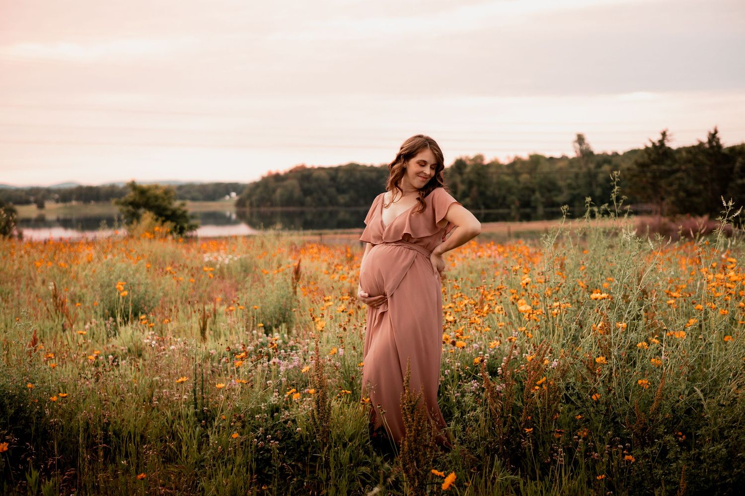 A pregnant woman in a pink dress walks through a vibrant field of yellow and orange wildflowers at sunset.