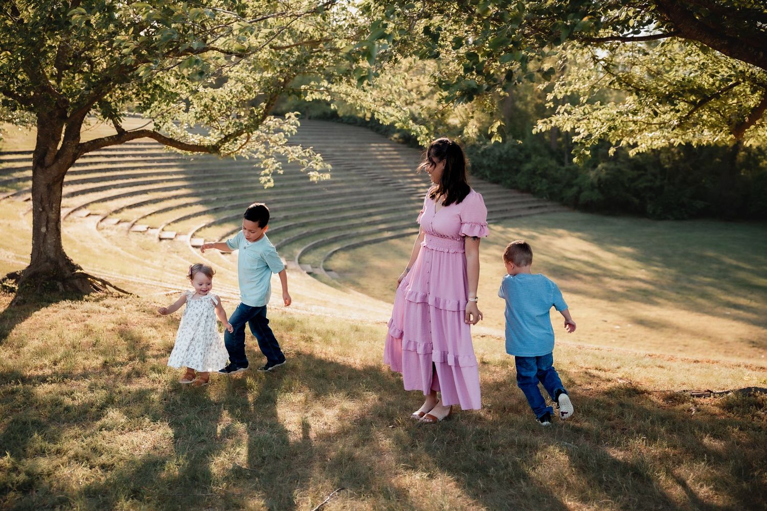 A mother in a pink dress walks with three young children on a grassy hill beneath a large tree in a park.