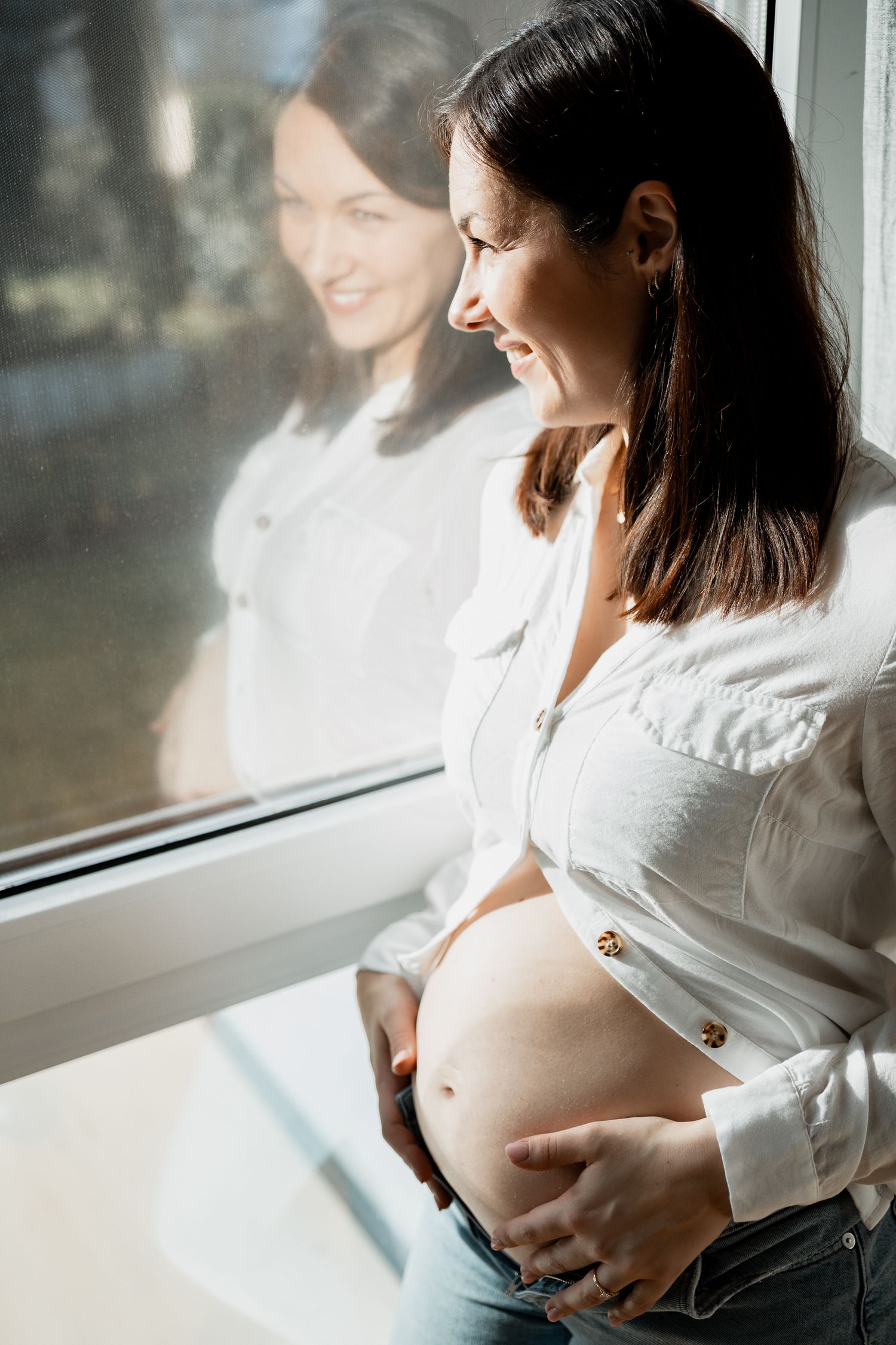Pregnant woman in white shirt sitting by window, holding belly in tender moment during maternity photoshoot series.