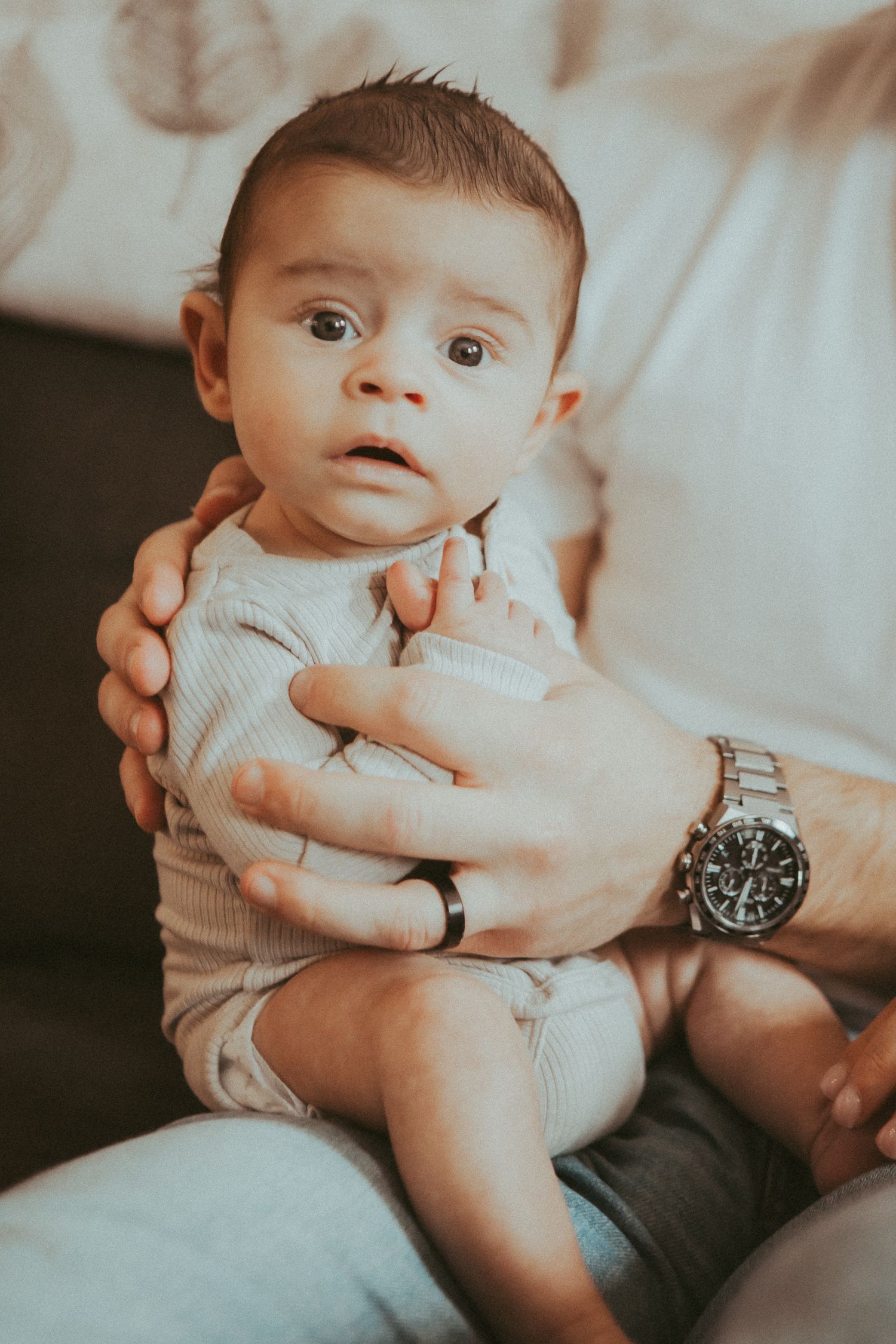 Baby in cream sweater looking up while being held by adult hands wearing watch.