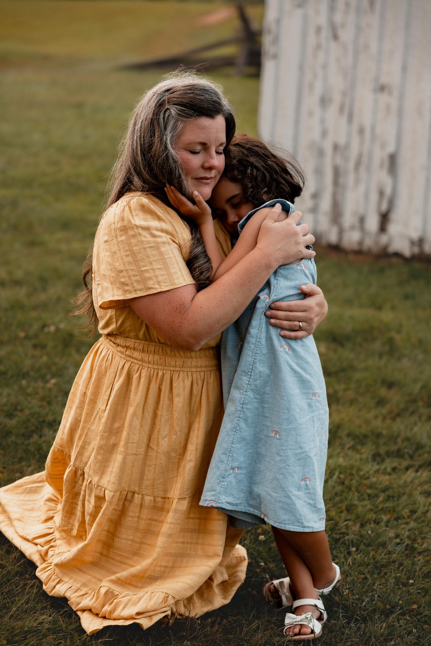 A mother in a yellow dress tenderly hugs her young daughter outdoors on a green lawn.