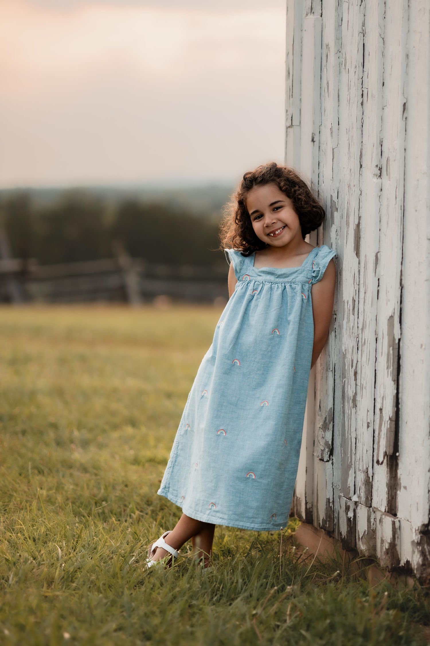 A smiling young girl in a light blue dress leans against a white barn wall in a green field at sunset.