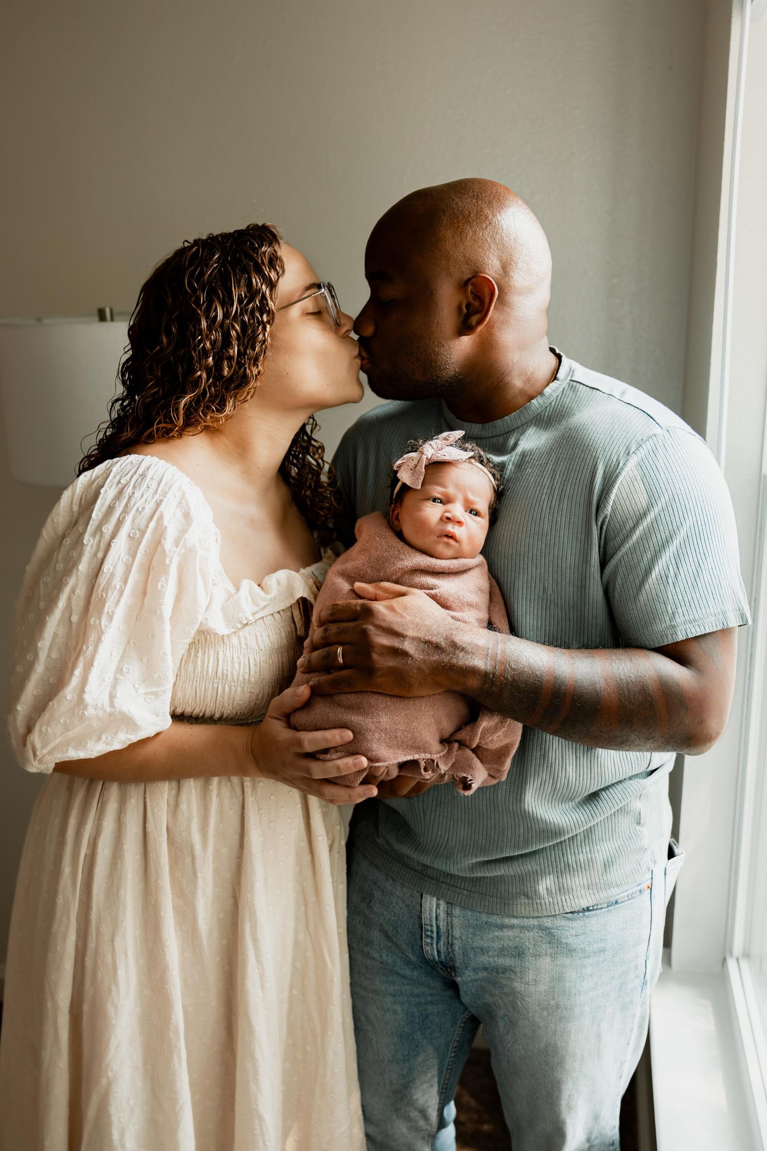 A couple shares a kiss while holding their swaddled newborn baby girl with a pink bow headband.