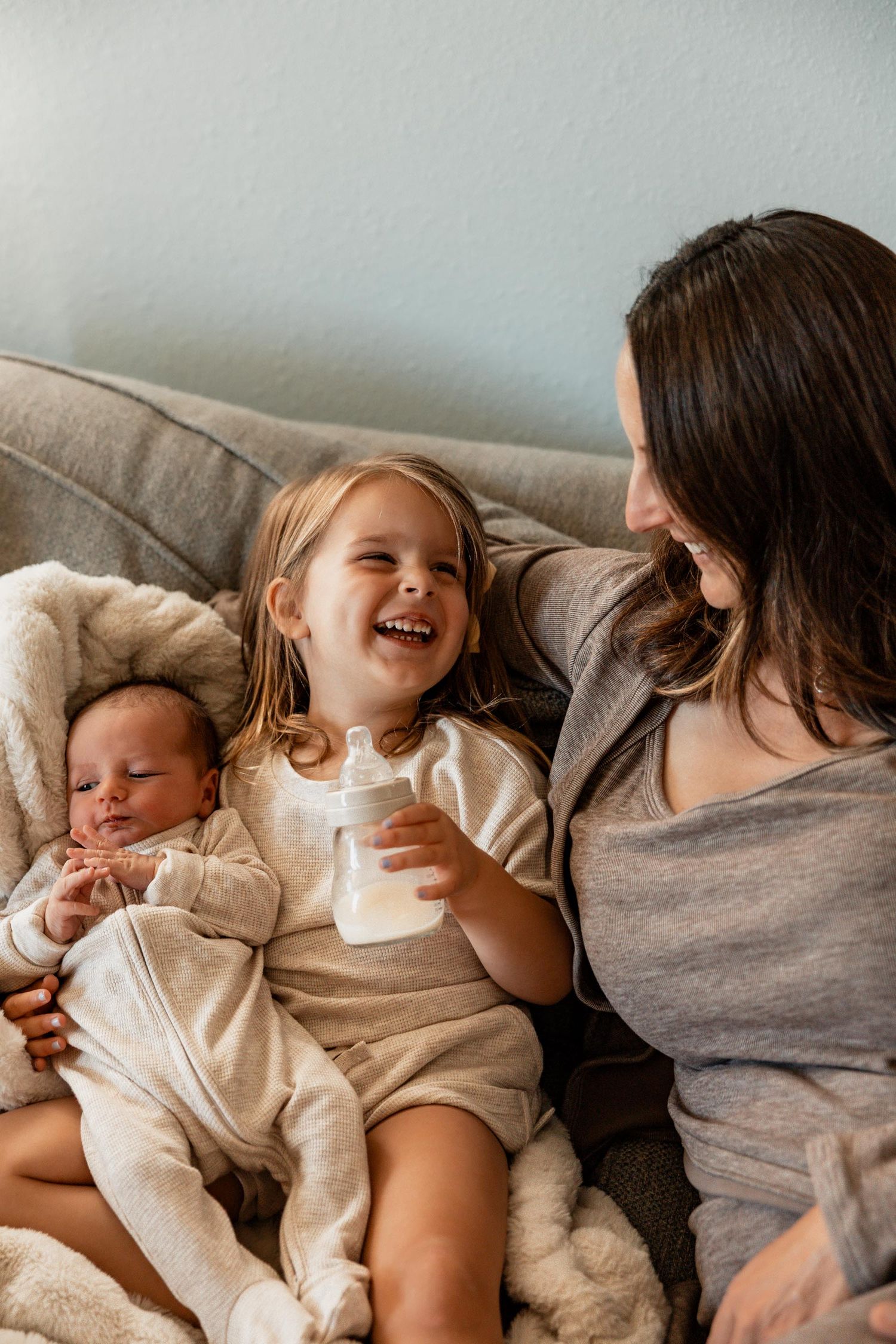 A mother and toddler girl laugh together on a couch while holding a baby bottle next to their sleeping newborn.