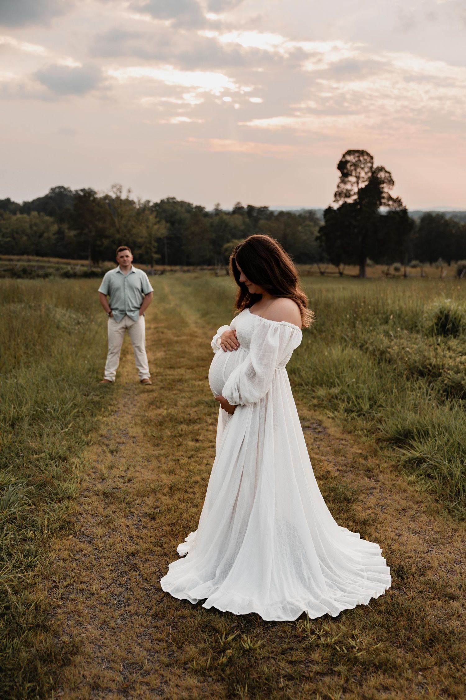 Pregnant woman in flowing white off-shoulder gown stands on a field path at sunset, man watching in the background.