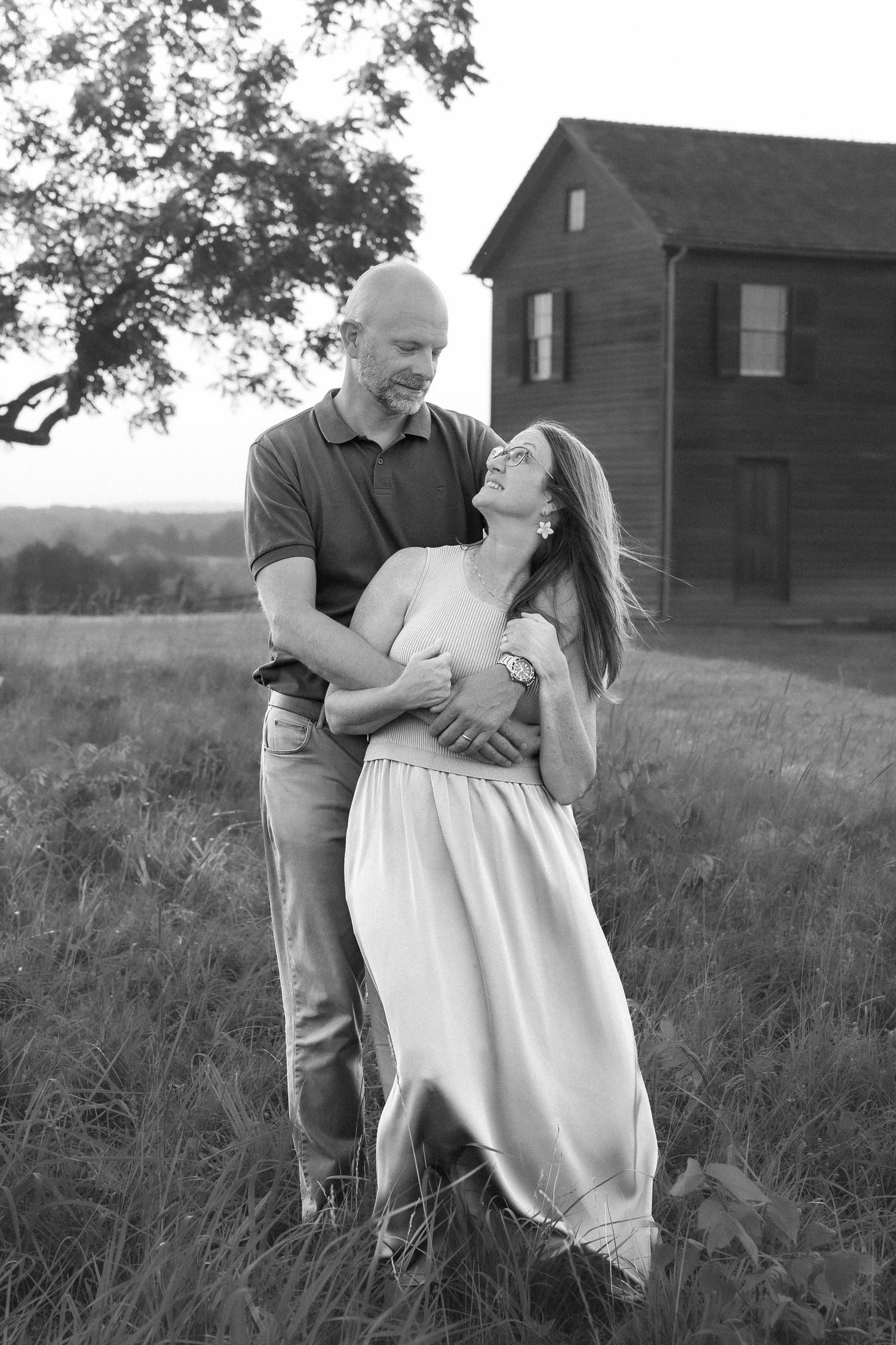 Black and white photo of a couple embracing and smiling in a field with a rustic barn in the background.