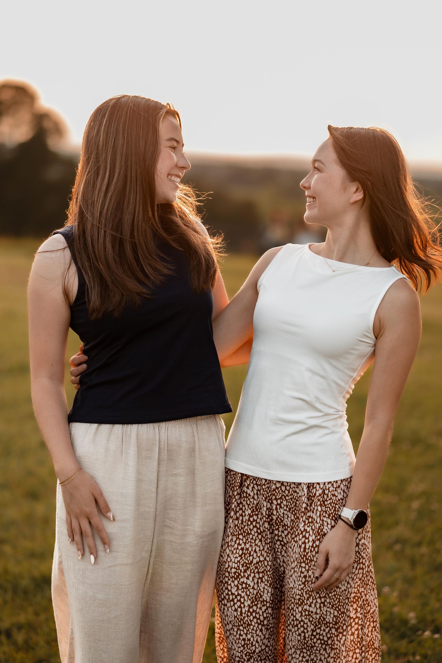 Two smiling redhead women in skirts enjoy a golden sunset outdoors, sharing a warm moment together.