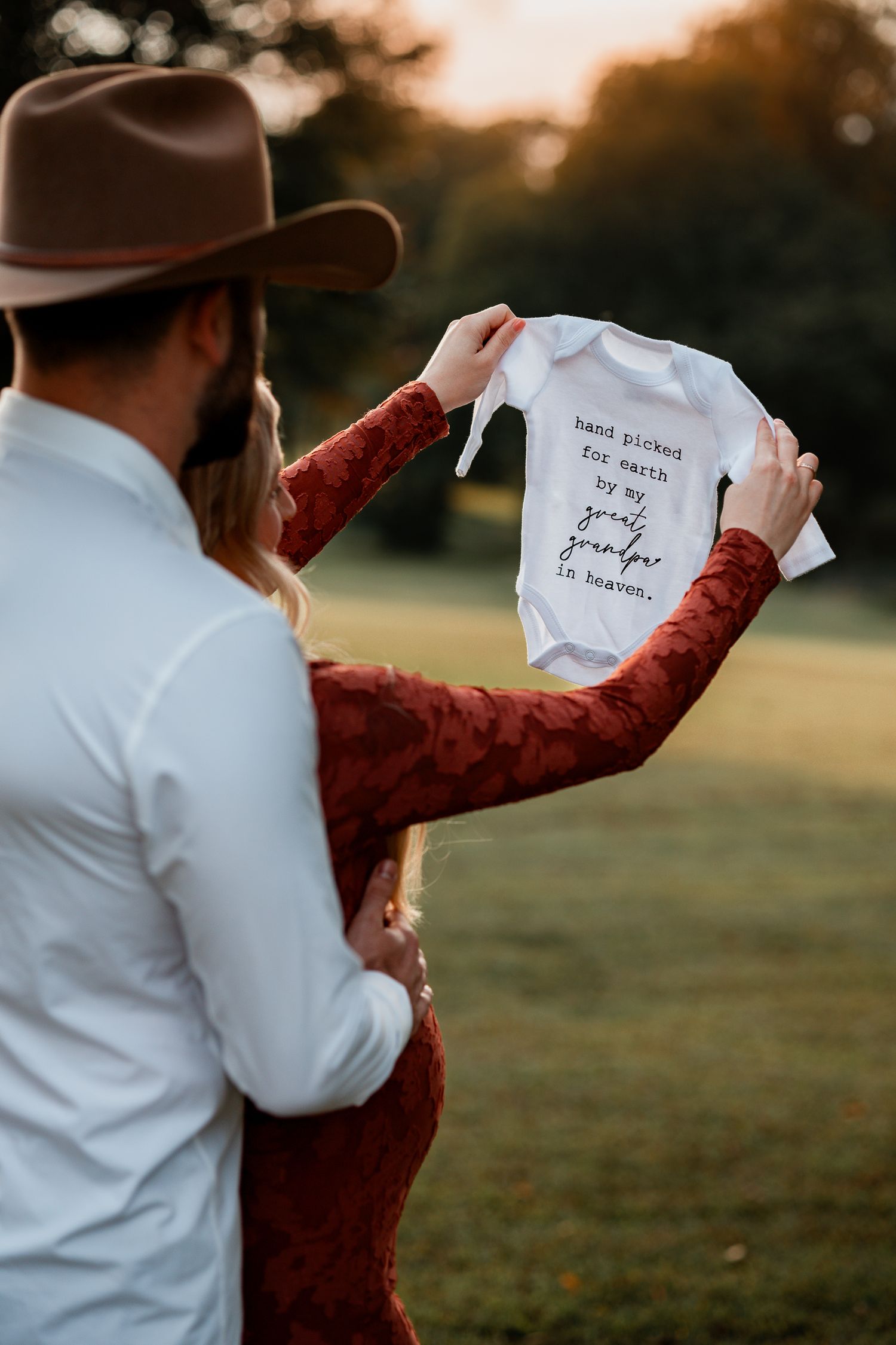 A couple holds up a small white baby onesie in a sunset field setting.