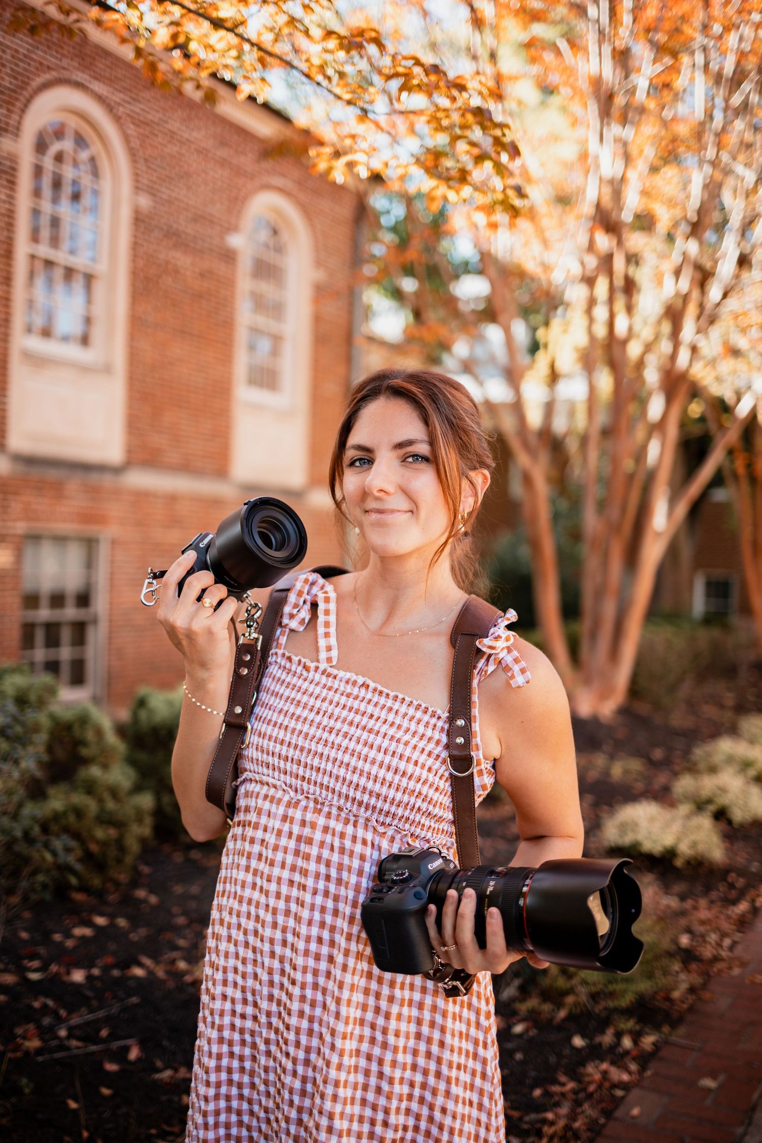 A photographer in a red gingham dress holds a professional camera while standing near a brick building in autumn.