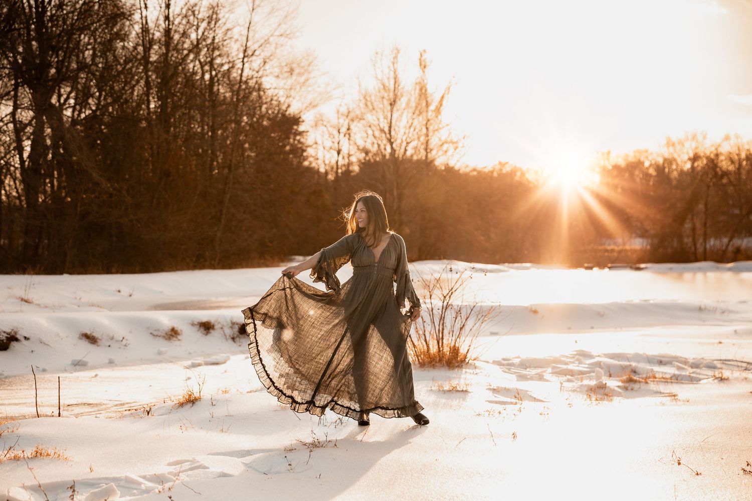 A person in a flowing dress stands in snowy field at sunset while sunlight creates a warm golden glow through bare trees.