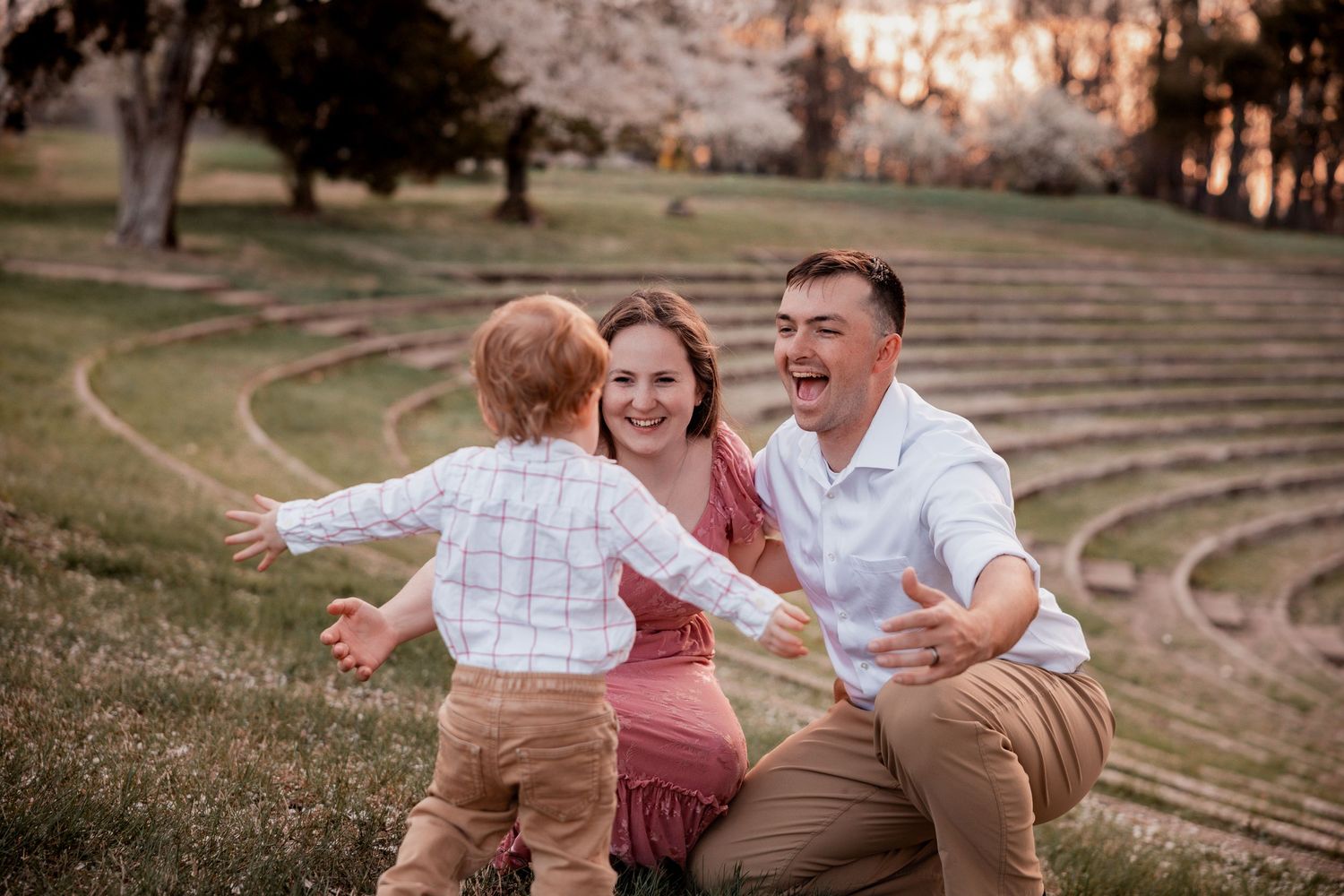 A joyful family moment outdoors as a toddler runs toward smiling parents sitting on green grass near blooming trees.