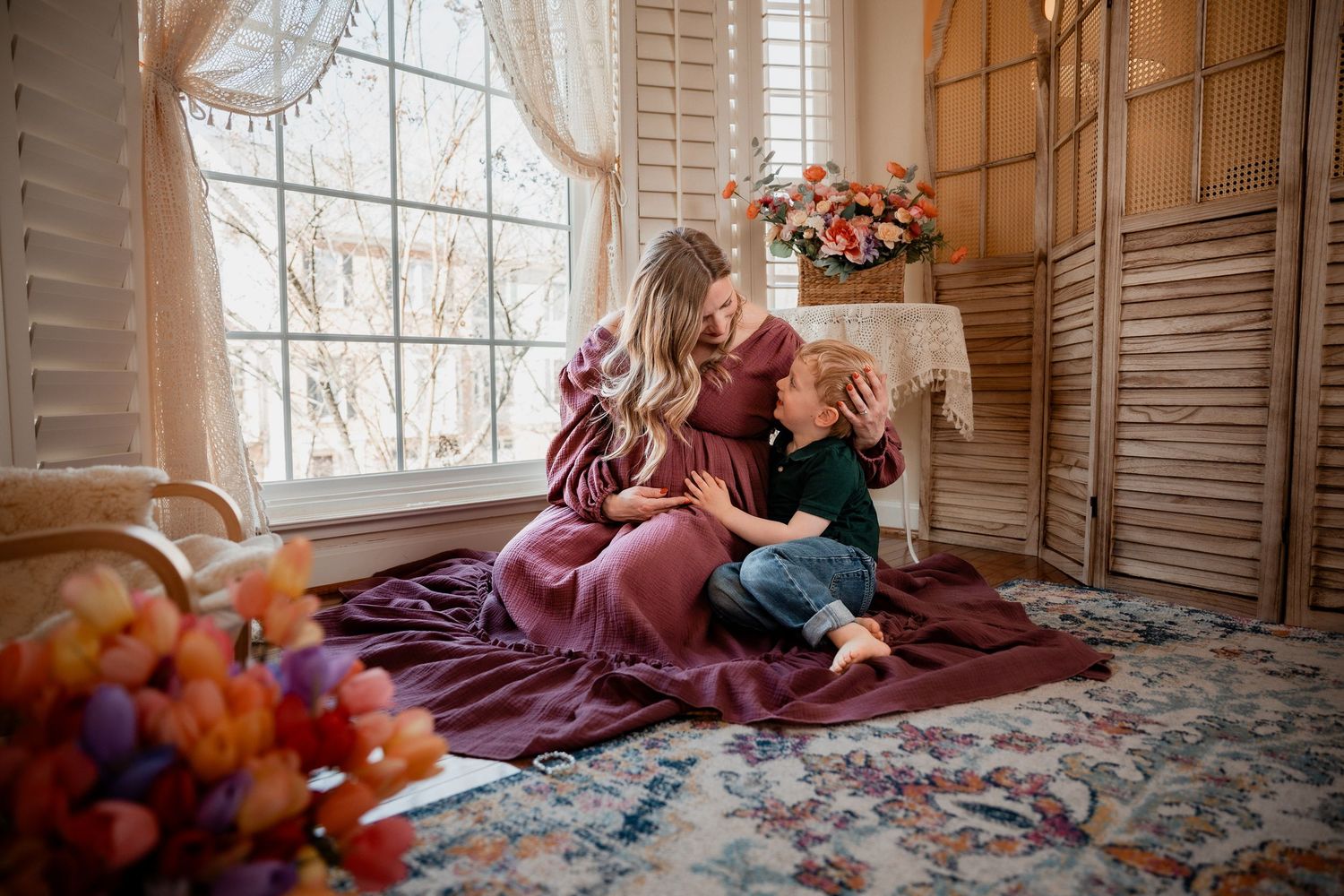 Pregnant woman in burgundy dress sitting on floor with young boy by large window with flowers.