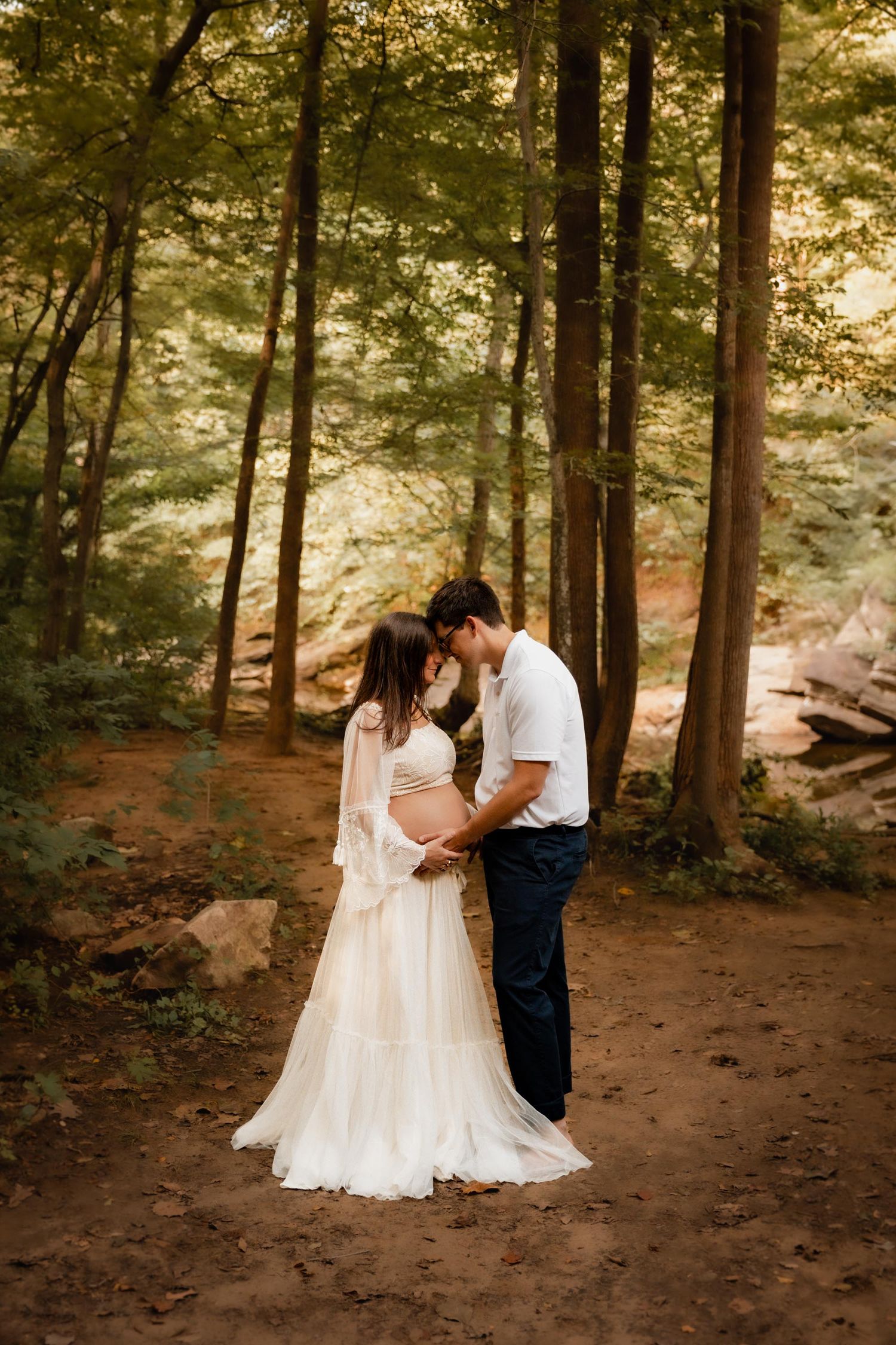 Couple sharing a tender kiss in a forest, woman in flowing white maternity gown, man in white shirt and dark pants.