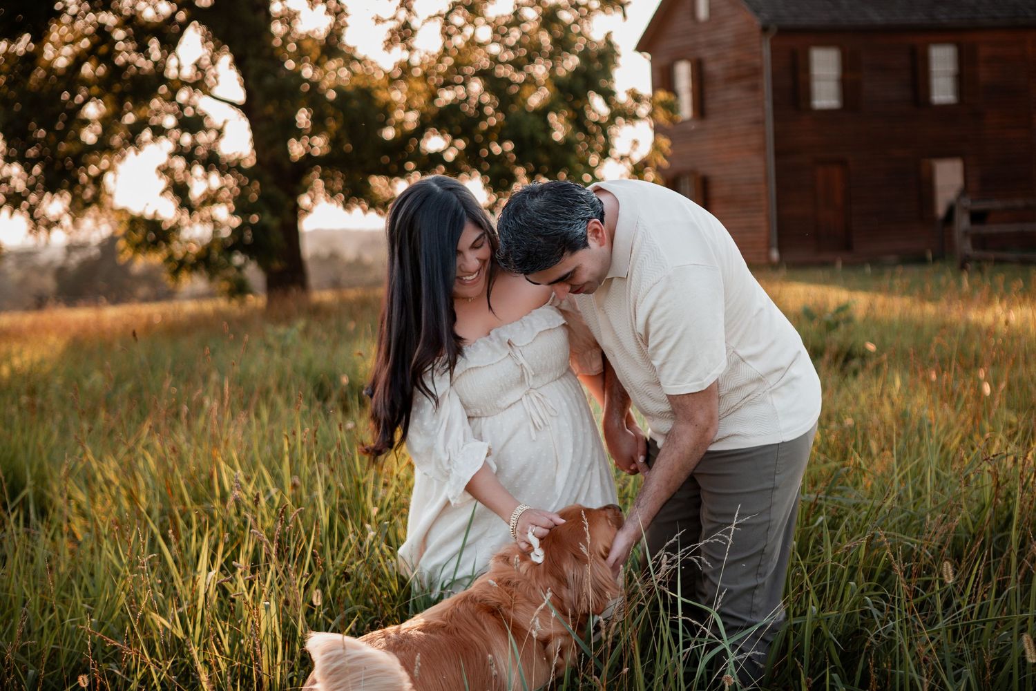 A couple in white outfits petting their golden retriever in a grassy field at sunset with a brick house in the background.
