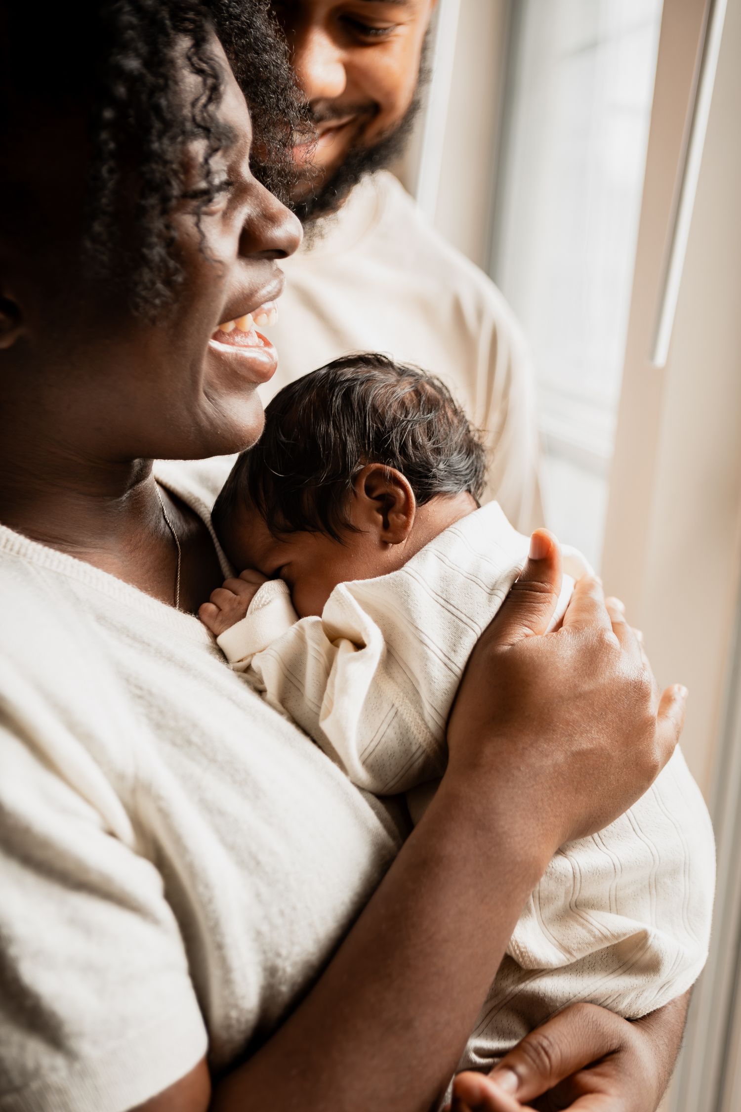 Parents hold their sleeping newborn baby next to a bright window in a tender family moment.