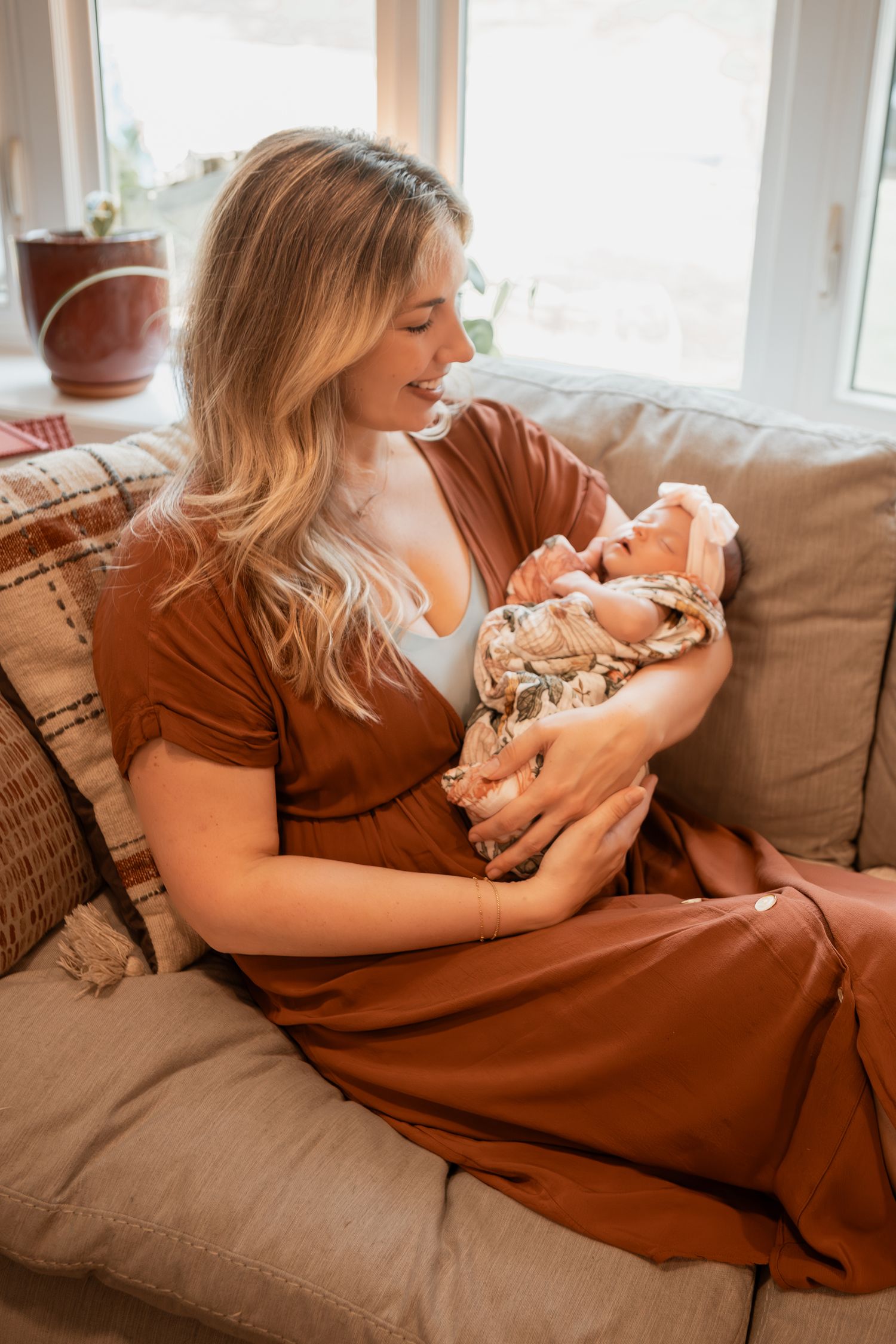 A woman in a brown dress sits on a gray couch while cradling a swaddled newborn baby in natural window light.