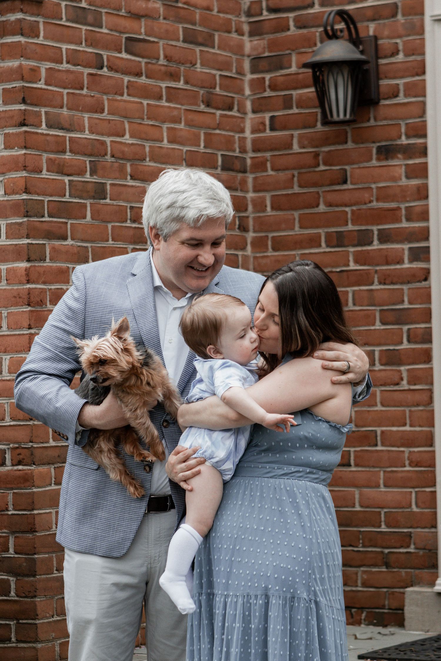 Family portrait with small dog against red brick wall showing affection and warm embrace.