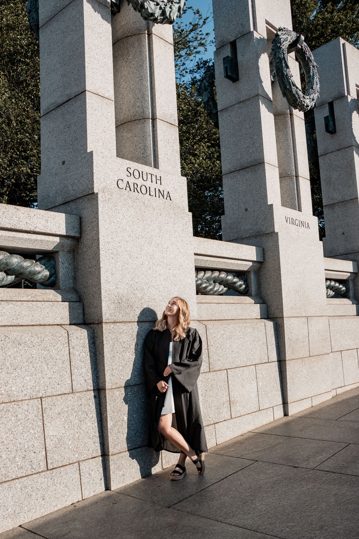 Graduate in dark gown poses against South Carolina pillar at memorial or monument.