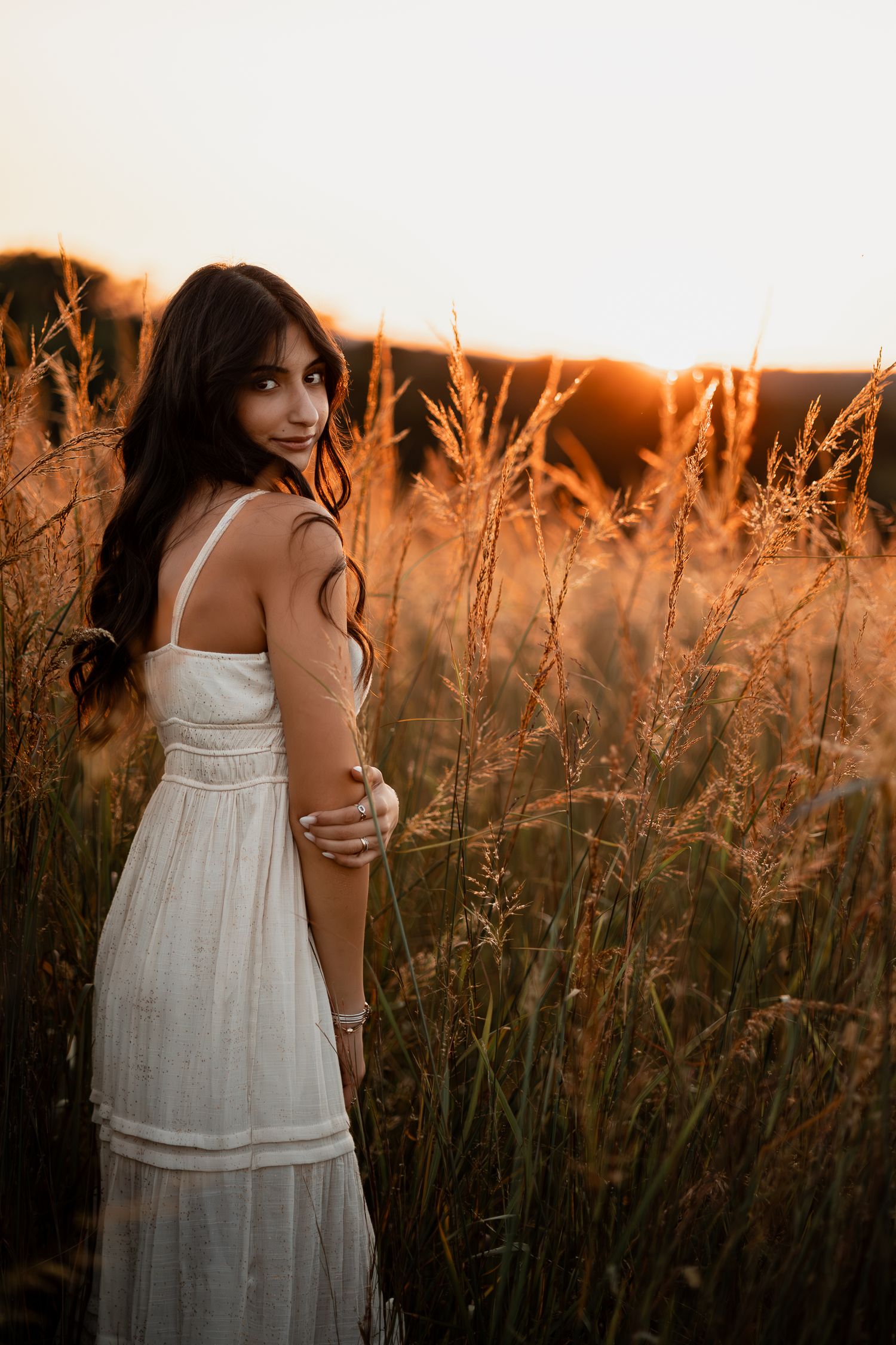 Person in flowing white dress stands among tall grasses at sunset creating a dreamy silhouette.